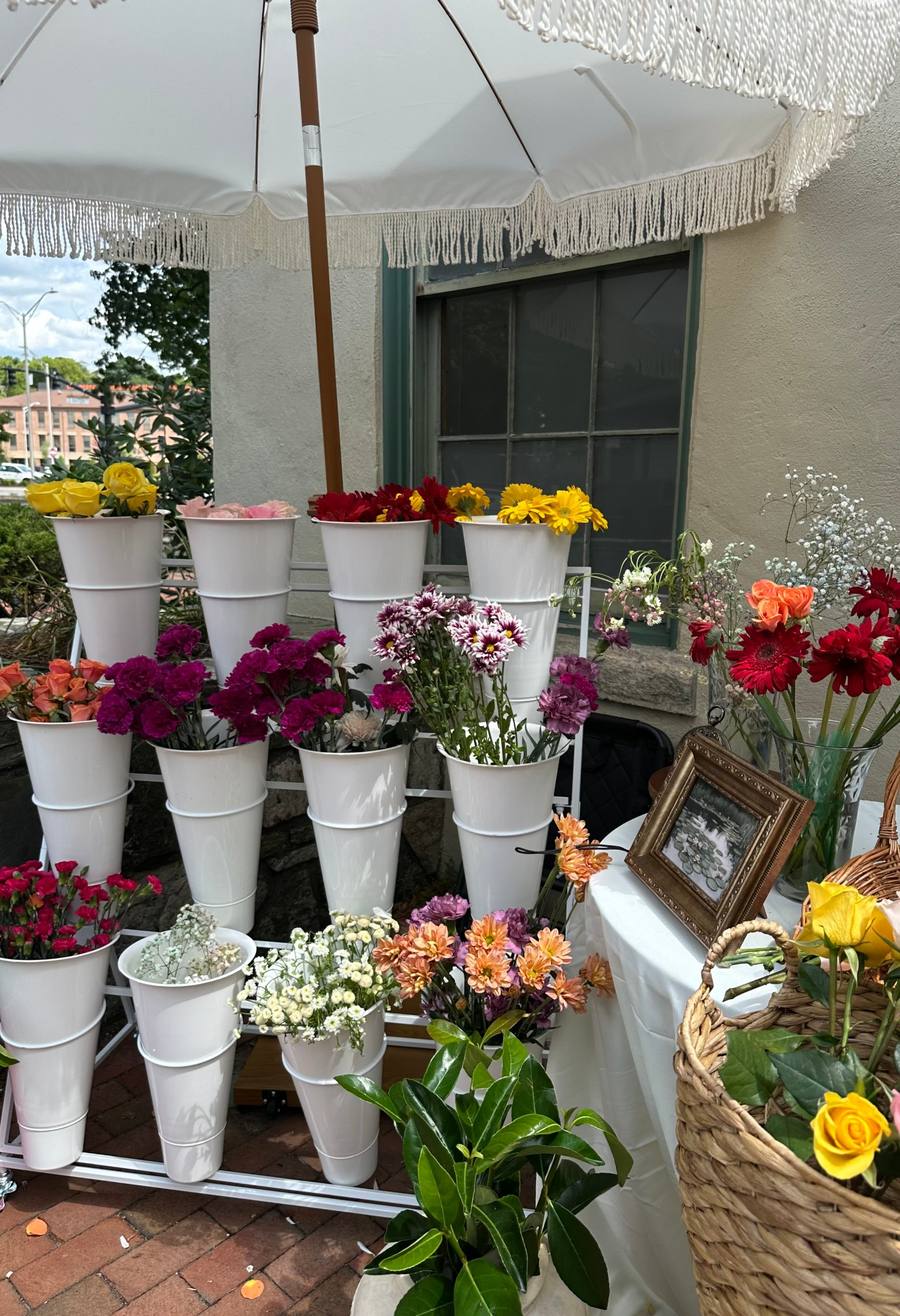 The Sunday Blooms cart under a white fringe umbrella, stocked with roses, gerberas, and carnations